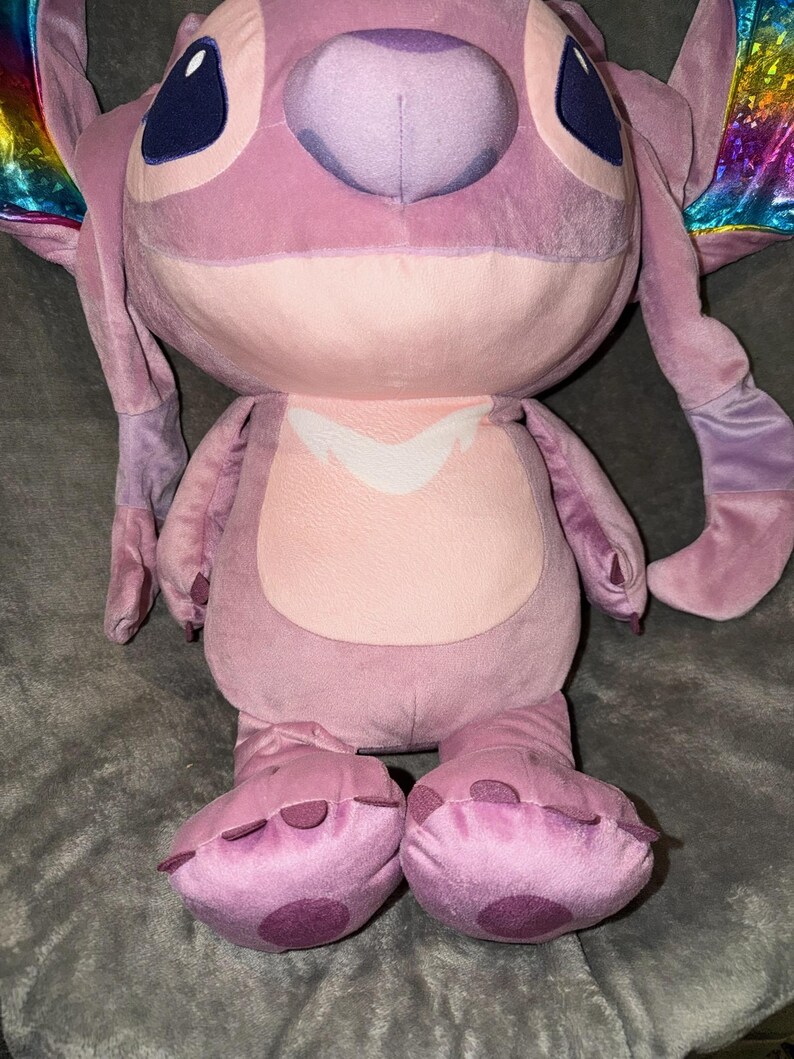Puede incluir: Una gran mu&ntilde;eca de peluche Angel en tonos rosas y morados. La mu&ntilde;eca tiene ojos azules grandes, orejas largas y un hocico redondeado. Los pies de la mu&ntilde;eca tienen manchas moradas oscuras. La mu&ntilde;eca tiene alas de colores arco&iacute;ris.