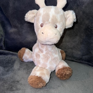 May include: A plush giraffe toy with a light brown and white spotted pattern. The giraffe has brown eyes, ears, and hooves. The stuffed animal is sitting on a gray surface.