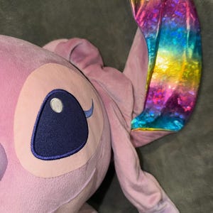 Puede incluir: Un peluche rosa claro de un personaje con ojos azules grandes y una oreja de color arco&iacute;ris. El juguete tiene una cabeza grande y orejas largas. El ojo del juguete es de color azul oscuro.