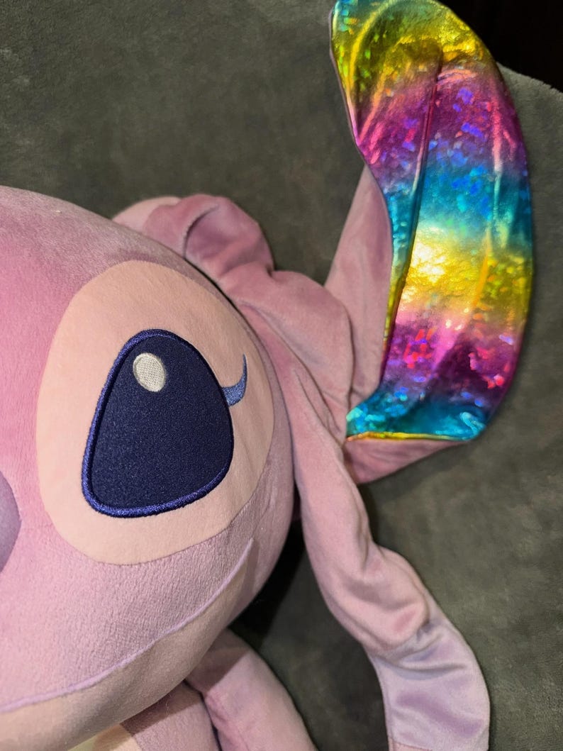 Puede incluir: Un peluche rosa claro de un personaje con ojos azules grandes y una oreja de color arco&iacute;ris. El juguete tiene una cabeza grande y orejas largas. El ojo del juguete es de color azul oscuro.
