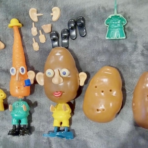 May include: A collection of plastic toy components including a brown potato-shaped body, an orange carrot-shaped body, two additional potato-shaped pieces, a green suit, a yellow suit, and various facial features like ears, eyes, and noses.