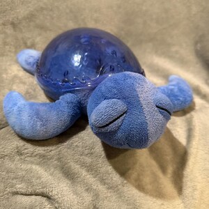 May include: A plush blue turtle toy with a clear, textured shell. The turtle has soft, light blue flippers and a head with closed eyes. The toy is lying on a gray, textured surface.