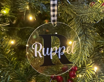 May include: Clear round Christmas bauble with a black and white checked ribbon. The bauble has a black letter "R" and the name "Ruppel" written in white.