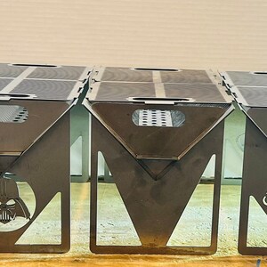 May include: Five metal fire pits with different team logos. The logos are the Raiders, Darth Vader, the Seahawks, and the Cowboys. The fire pits are all black and have a rectangular shape.