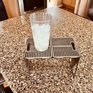 May include: A stainless steel folding table with a grid pattern on the top. The table is folded up and has a glass with ice and a clear liquid inside sitting on top of it.