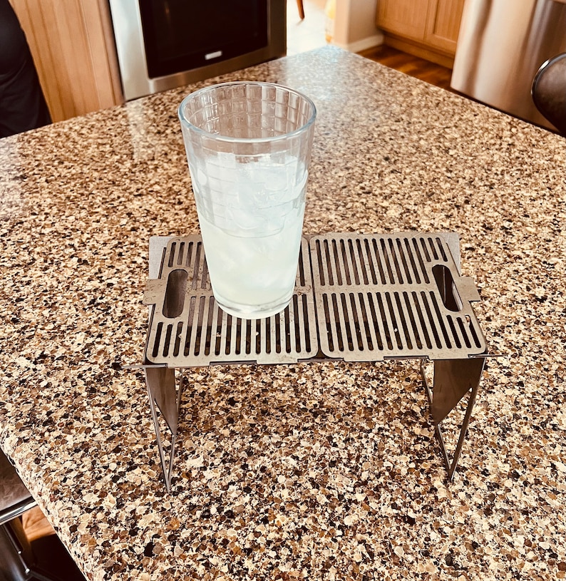 May include: A stainless steel folding table with a grid pattern on the top. The table is folded up and has a glass with ice and a clear liquid inside sitting on top of it.