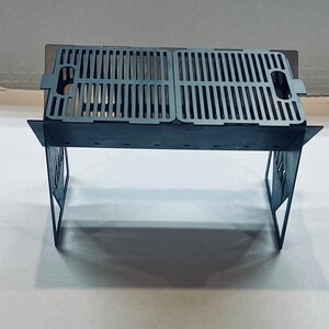 May include: A black metal folding grill with a rectangular grilling surface. The grill is made of sturdy metal and has a simple, functional design.