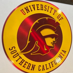 May include: A round, metallic sign featuring the words "UNIVERSITY OF SOUTHERN CALIFORNIA" in yellow lettering. The center design is a red Trojan warrior head, also in a metallic finish. The sign has a yellow border.