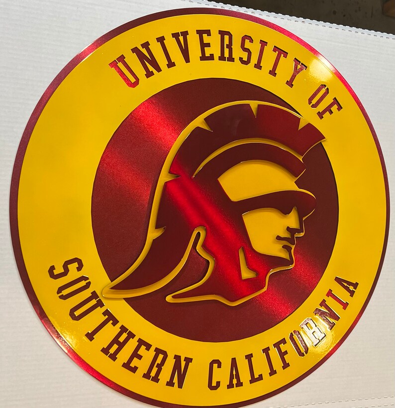May include: A round, metallic sign featuring the words "UNIVERSITY OF SOUTHERN CALIFORNIA" in yellow lettering. The center design is a red Trojan warrior head, also in a metallic finish. The sign has a yellow border.