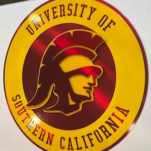 May include: A circular sign featuring the University of Southern California logo. The sign has a yellow outer ring and a maroon inner circle. The logo includes a Trojan warrior head in yellow and red, with the university name in maroon.