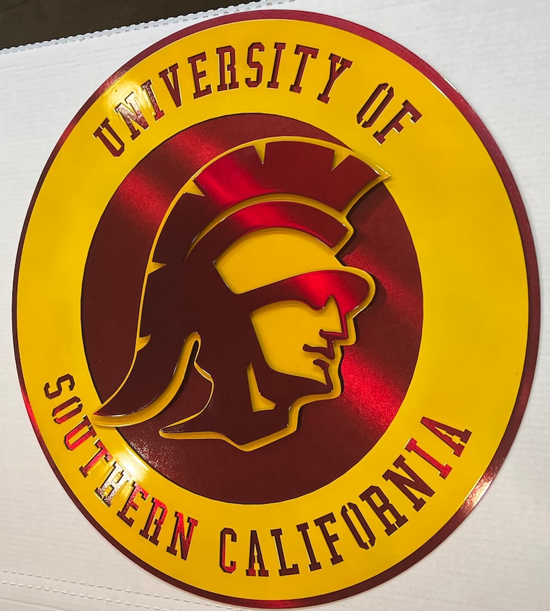 May include: A circular sign featuring the University of Southern California logo. The sign has a yellow outer ring and a maroon inner circle. The logo includes a Trojan warrior head in yellow and red, with the university name in maroon.