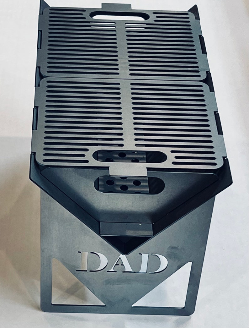 May include: A black metal fire pit with a grill grate and the word "DAD" engraved on the front.