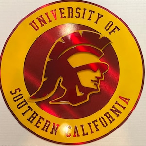 May include: A round, metallic sign featuring the University of Southern California logo. The design includes a Trojan warrior profile in maroon, set against a red and yellow background. The university name is displayed in a yellow, outlined font.