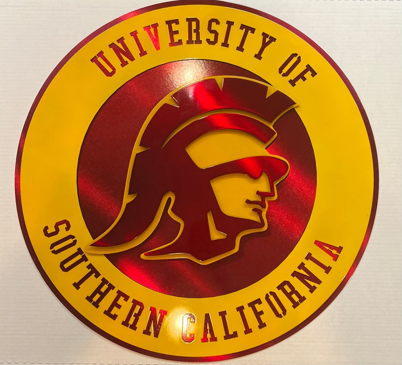 May include: A round, metallic sign featuring the University of Southern California logo. The design includes a Trojan warrior profile in maroon, set against a red and yellow background. The university name is displayed in a yellow, outlined font.