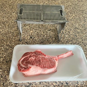 May include: A raw tomahawk steak on a white styrofoam tray with a clear plastic wrap. A stainless steel grill with a folding stand is in the background.