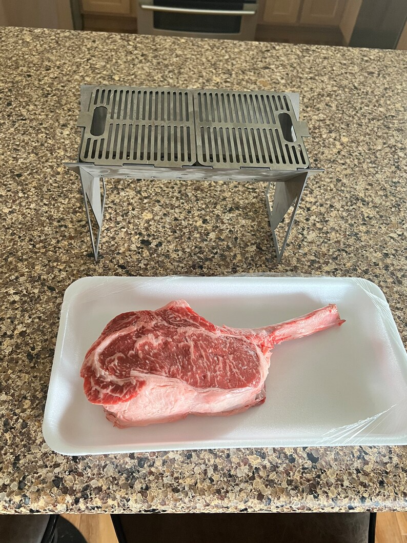 May include: A raw tomahawk steak on a white styrofoam tray with a clear plastic wrap. A stainless steel grill with a folding stand is in the background.
