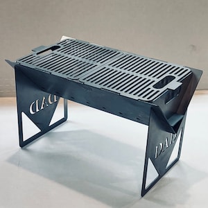 May include: A portable, stainless steel grill with a folding design. The grill has a rectangular cooking surface with a grid pattern and two handles. The grill is supported by two triangular legs with the words "CIAC" and "DAD" cut out.
