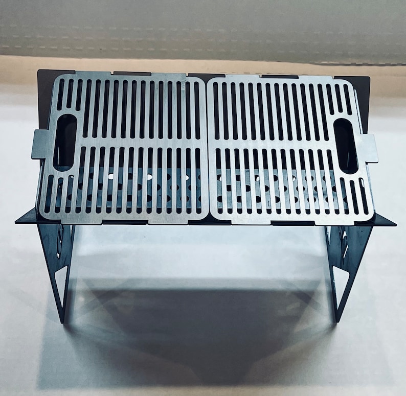 May include: A silver folding grill with a rectangular grilling surface. The grill is made of metal and has a simple design.