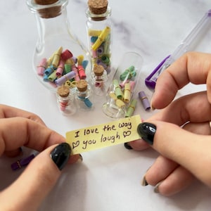 Könnte beinhalten: Mehrere kleine Glasflaschen mit Korken, gefüllt mit bunten, aufgerollten Papiernotizen. Ein gelber Haftzettel mit der Aufschrift "I love the way you laugh" mit Herzsymbolen. Ein Stift und kleine Wäscheklammern sind ebenfalls zu sehen.