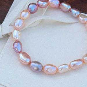 May include: A bracelet of iridescent freshwater pearls in shades of pink, peach, and lavender. The oval-shaped pearls are strung together, resting on a white fabric pouch against a dark wood surface.