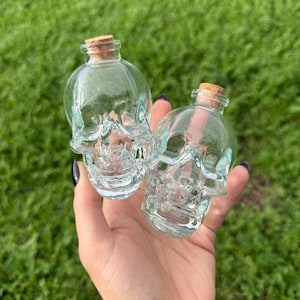 May include: Two clear glass skull-shaped bottles with cork stoppers. The bottles are held in a hand against a backdrop of green grass. The skull design is detailed, and the bottles appear to be small decorative items.