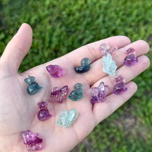 May include: A collection of small, colorful crystal figurines in various shapes, including ducks, crowns, and other abstract forms. The crystals are shades of purple, green, and blue, held in a hand against a green background.
