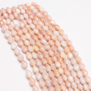 May include: A collection of faceted, teardrop-shaped peach-colored beads arranged in multiple parallel vertical strands. Each bead features a polished, multi-faceted surface that reflects light, creating a subtle shimmer across the light orange and cream-toned gemstone-like material.