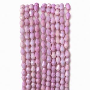May include: A collection of six vertical strands of faceted, pear-shaped beads in a soft, iridescent light pink hue. Each bead features a polished, multi-faceted surface that reflects light, arranged in uniform rows on thin, light-colored stringing material.