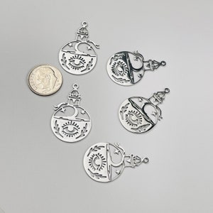 May include: Five silver-toned metal charms shaped like potion bottles, each featuring a cutout design of an eye, a crescent moon, stars, and leafy sprigs. A United States dime is included for scale, measuring approximately 0.7 inches in diameter.