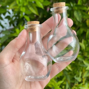 Puede incluir: Dos pequeñas botellas de vidrio transparente con tapones de corcho. Las botellas tienen forma redondeada y cuello estrecho. El fondo es una vista borrosa de follaje verde.