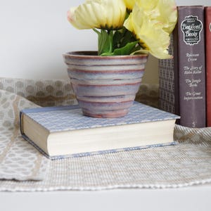 Op de afbeelding: Een keramische plantenpot met een gestreept patroon in tinten paars en bruin, met een boeket gele bloemen erin. De pot staat op een stapel boeken met de titels "Best Loved Books" op de ruggen.