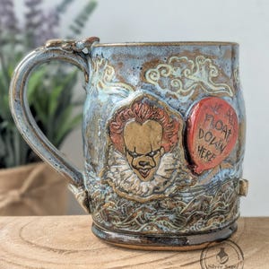 May include: Handmade ceramic mug with a blue and brown glaze. The mug features a relief of a clown with red hair and a red balloon with the text "WE ALL FLOAT DOWN HERE". The handle has a decorative design.