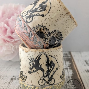 May include: Two stacked ceramic cups with a speckled cream glaze. Each cup features a black ink illustration of a bear wearing a crown and riding a bicycle, accompanied by sunflower motifs and a textured, carved leaf pattern around the base.