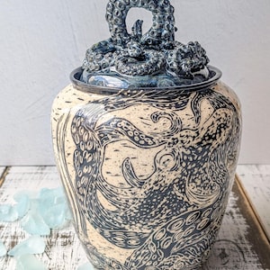 May include: A ceramic jar with a cream-colored body featuring black octopus illustrations and a dark blue lid topped with a sculpted octopus tentacle handle. The base of the jar displays a circular logo with the text Silver Sage Ceramics.