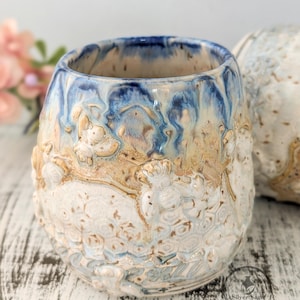 May include: A ceramic tumbler featuring a relief pattern of bees and honeycomb. The upper rim is glazed in deep blue, transitioning into a sandy tan and white base. A circular logo with the text Silver Sage Ceramics is visible on the lower right.