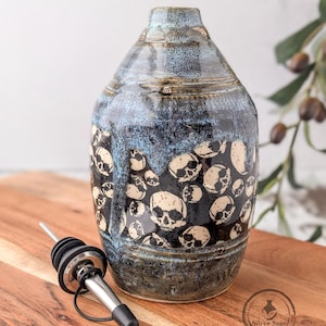 May include: A ceramic oil cruet featuring a blue and black speckled glaze with a pattern of cream-colored skulls. The bottle has a narrow neck and includes a stainless steel pourer spout with a black rubber stopper and a small black cap.