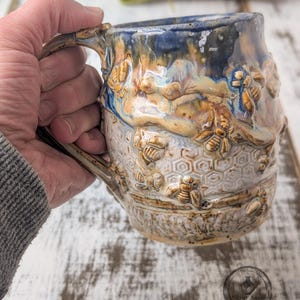 May include: A handmade ceramic mug with a bee and honeycomb design. The mug features a blue, tan, and white glaze, with sculpted bees and honeycomb patterns. The handle is integrated into the design. A unique piece for coffee or tea.