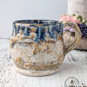 May include: A handcrafted ceramic mug with a textured surface and a unique design. The mug features a blue and brown glaze on the rim, with a beige and brown honeycomb pattern on the body. The handle is also glazed in shades of blue and brown.