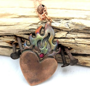 May include: A copper-toned heart pendant with a flame design, featuring a small red gemstone. The pendant hangs from a delicate copper chain. The flame has iridescent colors of green, blue, and orange.