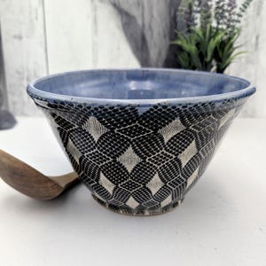 May include: A ceramic bowl with a blue interior and a black and white geometric pattern on the exterior. The bowl has a wide, open shape and sits on a white surface. A wooden spoon rests nearby.