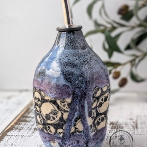 May include: A ceramic oil bottle with a stainless steel spout. The bottle features a blue, purple, and white glaze with a skull pattern. The bottle has a rounded shape and a narrow neck. The Silver Sage Ceramics logo is visible.