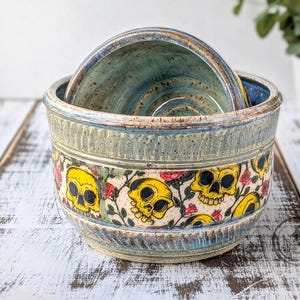May include: A set of three ceramic bowls. The largest bowl is light blue with a yellow skull and floral design around the exterior. The two smaller bowls are nested inside the larger bowl and are a darker blue.