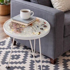 Puede incluir: Una pequeña mesa auxiliar redonda blanca con tres patas de horquilla. La mesa presenta un diseño de resina transparente con flores secas incrustadas, copos de oro y objetos decorativos en miniatura. Una taza de café blanca está sobre libros.