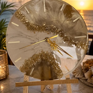 May include: A circular wall clock featuring a swirling blend of white and metallic gold resin. A diagonal band of textured gold glass crystals crosses the face. The clock has gold-colored hands and simple gold hour markers, displayed on a wooden easel.