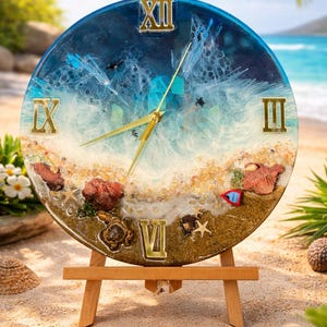 Puede incluir: Un reloj redondo con un diseño de playa. La esfera del reloj presenta una escena oceánica azul y blanca con números romanos y manecillas doradas. Se muestra sobre un pequeño caballete de madera, con un fondo de playa arenosa.