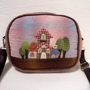 Puede incluir: Bolso bandolera de pana con un panel frontal acolchado con una escena de casas y árboles en aplique. El bolso tiene una base y un ribete marrones, un fondo de cielo colorido y una correa ajustable marrón.