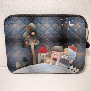 Pode incluir: Estojo de tecido acolchoado com uma cena de vila caprichosa. O design apresenta uma árvore com detalhes vermelhos, casas coloridas, uma lua crescente e estrelas em um fundo acolchoado azul e cinza. O estojo tem uma borda xadrez preta e cinza e um zíper verde.