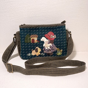 May include: A teal quilted crossbody bag with a whimsical appliqué design. The design features a girl in a red hat holding a bunny, a dog, and a house. The bag has a brown plaid strap and trim, and a zipper closure.
