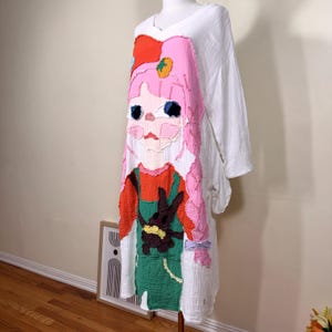 May include: White, long-sleeved dress with a large, colorful applique of a cartoon girl. The applique features pink hair, a red hat, and green overalls. The dress is displayed on a mannequin.