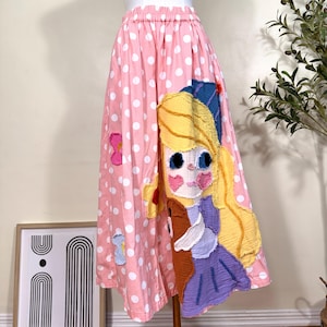 May include: A pink skirt with a white polka dot pattern featuring a large, textured fabric appliqué of a character with long yellow hair, blue eyes, and a purple dress, accented by smaller floral fabric patches in pink, yellow, and blue.
